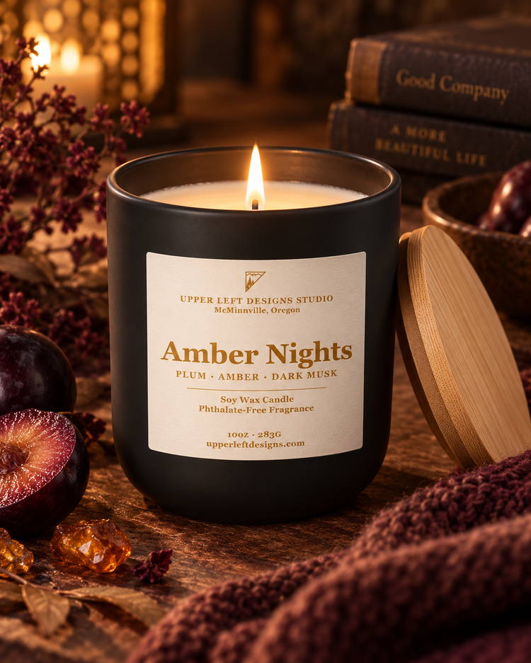 A lit black soy wax candle jar labeled 'Amber Nights' by Upper Left Designs Studio, styled on a wooden surface with plums, amber crystals, dried flowers, and a wooden lid nearby.