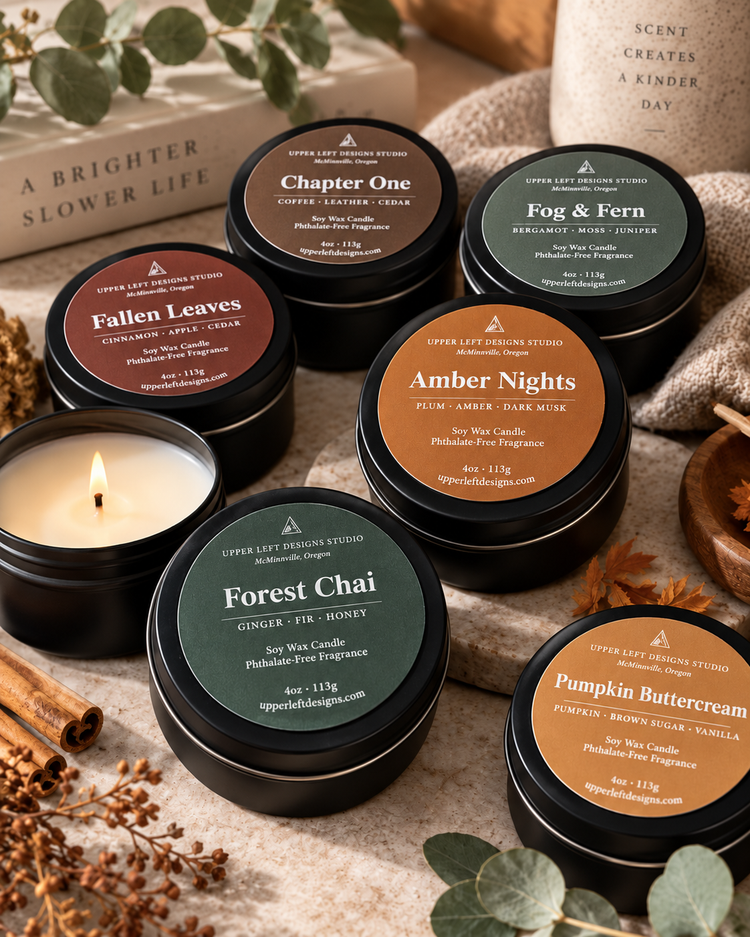 Six scented soy wax candles in round black metal tins with colorful labels, arranged on a neutral fabric surface with dried botanicals, cinnamon sticks, and a lit candle in a glass holder. Visible labels include Chapter One, Fog & Fern, Fallen Leaves, Amber Nights, Forest Chai, and Pumpkin Buttercream.